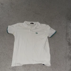 Vit pikétröja från Fred Perry - Vit pikétröja från Fred Perry med klassisk krage och knappar framtill. Tröjan har gröna och blå ränder på ärmsluten samt den ikoniska Fred Perry-loggan broderad på bröstet. Perfekt för en clean och sportig stil.