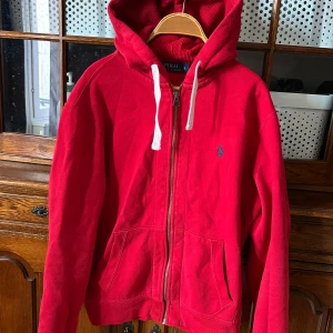 Röd hoodie från Polo Ralph Lauren - Riktigt snygg röd hoodie från Polo Ralph Lauren med dragkedja och vit snörning i huvan. Klassisk blå logga broderad på bröstet och två fickor framtill. Perfekt för dig som gillar en sportig och avslappnad stil.