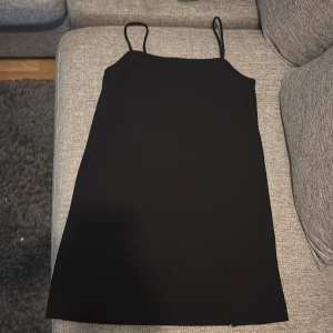 Svart kort klänning från H&M - Enkel svart klänning från H&M med smala axelband och rak passform. Klänningen är kort och har en liten slits framtill för extra detalj. Perfekt för dig som gillar minimalistisk stil och vill ha något som funkar till många olika looks.