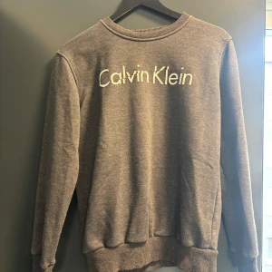 Grå sweatshirt från Calvin Klein Jeans - Snygg grå sweatshirt från Calvin Klein Jeans med vit logga framtill. Klassisk rund halsringning och ribbade muddar vid ärmslut och nederkant. Perfekt för en chill och stilren look. Lite nopprig 