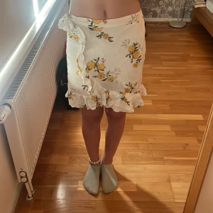 Vit blommig kjol från Lindex - Supersöt vit blommig kjol från Lindex med gult blommönster och volangkant. Kjolen har knyt i sidan och är gjord i ett lätt bomullstyg. Skriv till mig privat om ni vill ha en bättre bild
