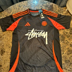 Colombia x Stüssy x Adidas fotbollströja L - Snygg svart och röd Colombia fotbollströja i samarbete mellan Adidas och Stüssy. Tröjan har korta ärmar, klassiska Adidas-ränder på axlarna, Stüssy-logga framtill och Colombia-märke. Tillverkad i lätt och ventilerande AEROREADY-material.