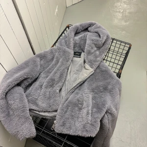 Grå teddyjacka från Urban Outfitters - Supermjuk och fluffig grå teddyjacka från Urban Outfitters med huva och dragkedja framtill. Jackan är croppad och har en riktigt cozy vibe, perfekt för kyliga dagar. Fodrad insida och två fickor framtill. Snygg att slänga över en hoodie eller t-shirt.