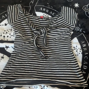Randig topp med knyt från Hell Bunny - Svart och vit randig topp från Hell Bunny med söt knytning framtill och korta ärmar. Toppen har en feminin passform och är gjord i mjukt material som känns skönt mot huden. Perfekt för dig som gillar en lite retro vibe och vill sticka ut med coola detaljer.