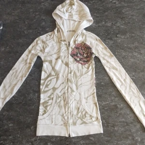 Vit hoodie med blommor och fjäril - Snygg vit hoodie med dragkedja och huva. Tröjan har ett unikt blommönster i beige och en stor röd blomma framtill. På ryggen finns en färgglad fjäril med rosdetaljer. Långärmad och med fickor framtill. Perfekt för dig som gillar statement-plagg!