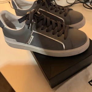  Balmain b court - Snygga grå sneakers från Balmain med vit sula och detaljer i mocka och skinn. Skorna har Balmain-logga på sidan och bak på sulan, samt mörkgrå snörning. Perfekt för dig som vill ha en clean och exklusiv look.