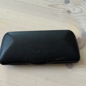 Svarta Balenciaga solglasögon med guldlogo - Snygga svarta solglasögon från Balenciaga med klassisk cat eye-form och guldiga BB-detaljer på skalmarna. Tillverkade i Italien och levereras i original svart fodral. Perfekta för dig som vill ha en lyxig och trendig accessoar.