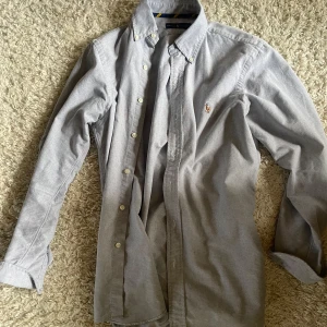 Grå skjorta från Ralph Lauren - Klassisk grå skjorta från Ralph Lauren med button-down krage och broderad logga på bröstet. Skjortan är långärmad och har vita knappar framtill. Perfekt för en clean och stilren look.