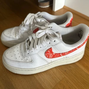 Nike Air Force 1 med röd paisley swoosh - Snygga vita Nike Air Force 1 sneakers med röd paisley-mönstrad swoosh och häl. Klassisk siluett med snörning och platt sula. Ovandel i skinn och detaljer i textil. Perfekta för dig som vill sticka ut med en unik touch på dina sneakers.