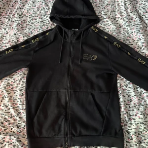 Svart EA7 hoodie med dragkedja - Svart hoodie från EA7 Emporio Armani med guldfärgade logotyper längs ärmarna och på bröstet. Tröjan har huva med snörning, hel dragkedja och två fickor framtill. Tillverkad i mjuk bomullsblandning, perfekt för en sportig och stilren look. Storlek M men passar som L