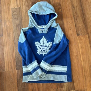 Toronto Maple Leafs blå hoodie NHL - Blå och grå hoodie med Toronto Maple Leafs-logga framtill. Tröjan har snörning vid halsen, huva och långa ärmar med gråa och blå ränder vid muddar och nederkant. Perfekt för dig som älskar hockey och vill visa ditt lagstöd.