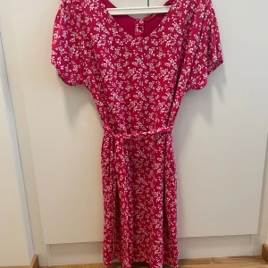 Rosa blommig klänning från s.Oliver - Supersöt rosa klänning från s.Oliver med vitt blommönster, v-ringning och knappar i ryggen. Modellen har korta ärmar, midja med matchande bälte och faller till under knäet. Perfekt för sommarens alla tillfällen!