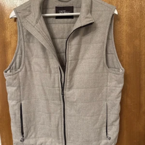 Ljusgrå quiltad dunväst Oscar Jacobson - Snygg ljusgrå quiltad dunväst från Oscar Jacobson med dragkedja framtill och två praktiska fickor med dragkedja. Västen har en stilren och enkel design med hög krage och raka linjer, perfekt att slänga över en hoodie eller skjorta.