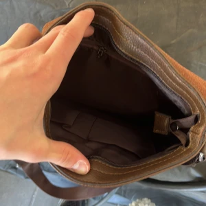 Brun axelväska från Tedikabolo - Snygg brun axelväska från Tedikabolo med schackrutigt mönster på framsidan och bred, justerbar axelrem. Väskan är gjord i skinnimitation och har flera fack, inklusive innerficka med dragkedja. Perfekt för att bära med sig allt viktigt i vardagen. OBS använd 5-6 gånger när jag reste utomlands förra året.