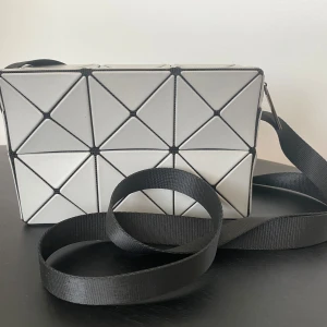 Vit axelväska från Bao Bao Issey Miyake - Säljer en unik vit axelväska från Bao Bao Issey Miyake med geometriskt 3D-mönster och svarta detaljer. Väskan har justerbar svart rem och dragkedja upptill. Perfekt för dig som gillar futuristisk design och vill sticka ut med en cool accessoar.