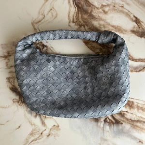 Silver axelväska från Bottega Veneta - Säljer en snygg silverfärgad axelväska från Bottega Veneta med klassiskt flätat mönster och robust handtag med knut. Väskan är gjord i skinn och har en guldfärgad dragkedja. Perfekt för dig som vill ha en stilren och trendig accessoar.