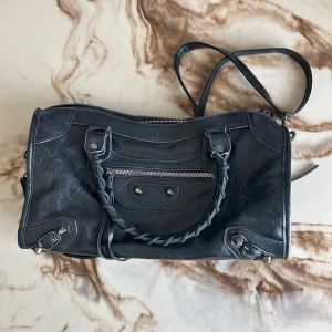 Svart Balenciaga axelväska - Säljer en svart axelväska från Balenciaga i skinn med flätat handtag, silverfärgade metalldetaljer och dragkedja både upptill och på framsidan. Väskan har både axelrem och handtag, samt flera fack för smart förvaring.