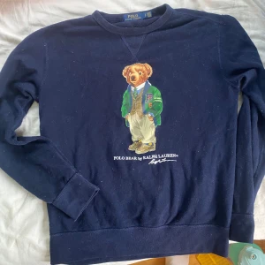 Mörkblå Polo Bear tröja Ralph Lauren - Mörkblå crewneck från Polo Ralph Lauren med ikoniska Polo Bear-trycket på bröstet. Tröjan har rund hals, långa ärmar och är gjord i mjuk bomull. Perfekt för dig som gillar klassisk preppy stil med en twist.