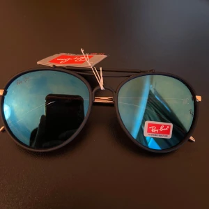 Ray-Ban svarta pilotbågar med blått glas - Snygga Ray-Ban solglasögon med klassisk pilotform. Svarta bågar i metall och spegelblå glas ger en cool vibe. Tunn dubbelbrygga och tunna skalmar med gulddetaljer. Perfekta för dig som vill sticka ut med stil.