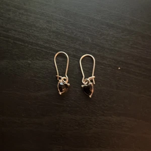 Silverörhängen med svarta hjärtan - Snygga örhängen i silverfärg med svarta hjärtformade stenar. Hängande modell med enkel krok och stilren design. Perfekta för dig som gillar en lite edgy men ändå söt look.