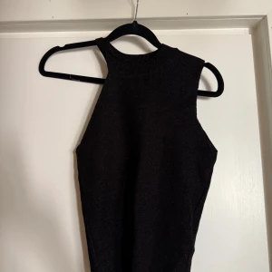 Svart glittrig ärmlös topp NA-KD - Snygg svart ärmlös topp från NA-KD med glittriga detaljer. Toppen har hög halsringning och är stickad i ett stretchigt material som sitter tight. Perfekt för dig som vill sticka ut lite extra med en stilren men ändå festlig look.