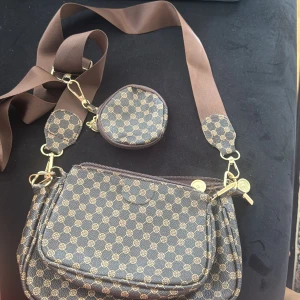 Brun axelväska med mönster och myntficka - Snygg brun axelväska med små cirkelmönster i beige och matchande bred axelrem. Väskan har guldfärgade detaljer och en tillhörande liten rund myntficka. Perfekt för att bära det viktigaste och ge outfiten en cool touch.