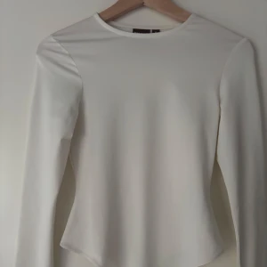 Vit långärmad bodysuit - Säljer en stilren vit långärmad bodysuit med åtsittande passform och spetsig nederkant. Perfekt att styla med jeans eller kjol för en clean look. Är från Biancas kollektion med Nelly.