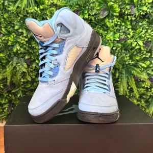 Nike Air Jordan 5 UNC blå sneakers - Nike Air Jordan 5 UNC sneakers i ljusblå mocka med svarta detaljer och silverfärgad plös. Skorna har klassisk snörning, meshpaneler på sidorna och broderad Jumpman-logga på plösen. Insidan har mönstrat foder och hälen pryds av en etikett med röd logga.