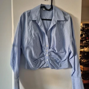 Blåvit randig skjorta från Zara - Snygg blå skjorta med vita ränder från Zara. Skjortan har croppad passform, långa ärmar med breda manschetter och coola knappar framtill. Materialet är mjukt och känns som bomull. Perfekt för dig som gillar trendiga detaljer och stilren look.
