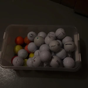 Golfbollar  - Bilderna visar en låda med golfbollar i olika färger och märken, samt en närbild på en vit Titleist-golfboll. Ingen bok syns på bilderna. Färgade 5 per boll ofärgade 3per boll