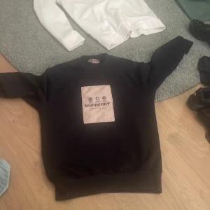 Svart Burberry sweatshirt med patch - Svart sweatshirt från Burberry med en beige patch framtill där loggan är tryckt i svart. Tröjan har rund halsringning och långa ärmar. Perfekt för dig som gillar stilrena och exklusiva plagg med tydlig logga. Betalning via Swish 