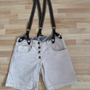 Beiga shorts med hängslen och knappar - Unika beiga/ljus rosa shorts med  hängslen och knappgylf. Shortsen har flera fickor både fram och bak samt snyggt randigt innerfoder i midjan. Perfekt för dig som vill sticka ut med en retrovibe och coola detaljer.Modellen på  tyget är skrynkligt, stretch. Midjan 82cm. Storlek lapp saknas, men den är storlek 38/40.