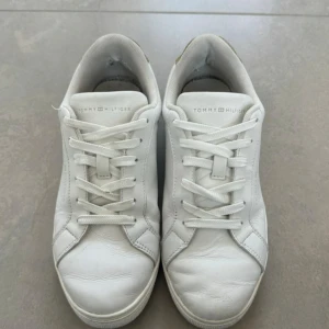 Vita sneakers Tommy Hilfiger guld - Snygga vita sneakers från Tommy Hilfiger med gulddetaljer. Klassisk låg modell med snörning. Perfekta för dig som gillar stilrena och trendiga skor med en touch av guld. Är använda men ändå bra skick ni ser på bilderna vart de är skråmor. Köpta för 1200 kr säljer för 250 kr.