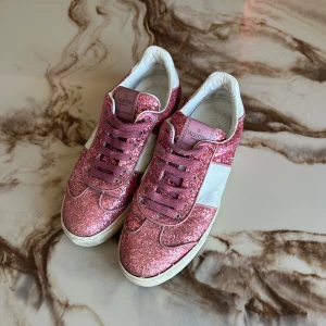 Valentino Garavani Flycrew glittriga - Snygga sneakers från Valentino Garavani i rosa glitter med vita detaljer och klassiska nitar baktill. Skorna har snörning, platt sula och är tillverkade i skinn med glittriga partier som verkligen sticker ut. Perfekta för dig som vill ha något extra till din outfit.