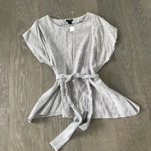 Grå ribbad blus med knytband  - Snygg ljusgrå ribbad blus från lindex med korta ärmar och rund halsringning. Toppen har ett matchande knytband i midjan som ger en fin siluett. Perfekt att styla med jeans eller kjol för en chill look.