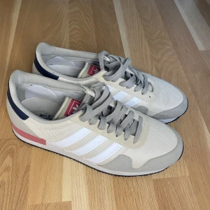 Adidas sneakers i beige och vitt - Snygga Adidas sneakers i beige textil med vita ränder och grå mockadetaljer. Skorna har grå snörning, svart hälkappa med röd till rosa Adidas-logga och rosa detalj på sulan. Klassisk retrostil som passar perfekt till streetwear. Skorna är bara använda en gång.