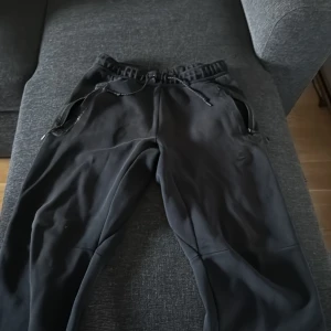 Svarta mjukisbyxor från Nike M - Svarta mjukisbyxor från Nike med dragsko i midjan och dragkedjor vid fickorna. Byxorna har en loose passform och är tillverkade i mjukt syntetmaterial. Perfekta för chill eller träning, med justerbara benslut för extra komfort.