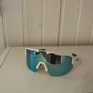 Bliz sportiga vita solglasögon - Snygga sportglasögon från Bliz med vit båge och spegelblått, stort linsformat glas. Perfekta för dig som gillar att sticka ut med en futuristisk vibe. Lätta och bekväma med svart detalj på skalmarna. Riktigt clean look för träning eller cykel.