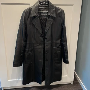 Svart lång skinnrock RockBoco - Snygg svart lång skinnrock från RockBoco med klassisk krage, stora fickor och knäppning framtill. Jackan har quiltat innerfoder och raka linjer som ger en clean och tidlös look. Perfekt för dig som vill sticka ut med en edgy vibe.