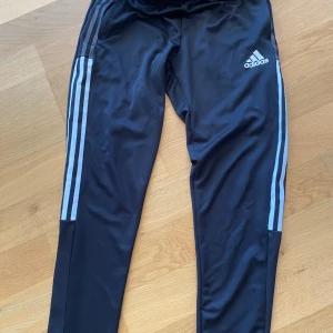 Svarta Adidas träningsbyxor M - Svarta träningsbyxor från Adidas med klassiska vita ränder längs sidorna och logga på låret. Byxorna har dragkedja vid benslut och är gjorda i polyester. Perfekta för träning eller chill, med slim passform och sportig look.