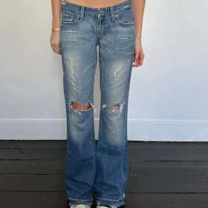 Blå slitna bootcut jeans - Snygga blå jeans med bootcut passform och slitna detaljer över knäna. Låg midja och klassisk femficksmodell. Jeansen har en avslappnad vibe med slitningar och ljusare partier framtill. Perfekta för en cool och avslappnad stil.
