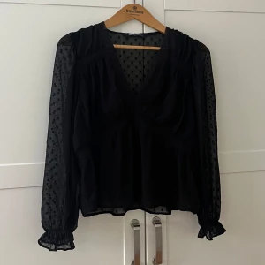 Svart blus med v-ringning från Zara - Supersnygg svart blus från Zara med v-ringning och transparenta ärmar i mesh med diskret rutigt mönster. Blusen har volang vid ärmslut och spetsdetaljer längs bysten. Perfekt för dig som gillar en chic och trendig look.