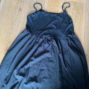 Svart kort klänning med smala axelband - Enkel svart klänning med smala axelband och utställd kjol. Överdelen är ribbad och har en tightare passform medan kjolen är mer flowy. Perfekt för dig som gillar minimalistisk stil och vill ha något bekvämt och snyggt.