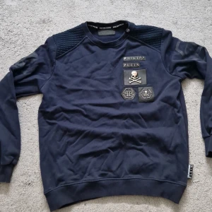 Mörkblå sweatshirt Philipp Plein - Snygg mörkblå sweatshirt från Philipp Plein med coola patchar och döskalle-detaljer på bröstet. Tröjan har quiltade axelpartier och långa ärmar. Perfekt för dig som gillar streetstyle och vill sticka ut lite extra.