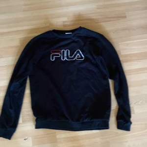 Svart Fila sweatshirt med logga - Svart sweatshirt från Fila med stor vit och röd logga på bröstet. Klassisk rund halsringning och långa ärmar. Mjuk och skön bomullsmix, perfekt för en chill och sportig stil. Kan förekomma katt hår för finns katt i huset 