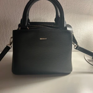 Svart handväska från DKNY  - Snygg svart handväska från DKNY i strukturerat skinn med guldfärgade detaljer och logotyp. Väskan har både handtag och avtagbar axelrem, Köpt från Zalando