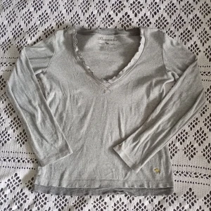 Grå tröja Abercrombie - Grå vintage supersöt långärmad tröja från Abercrombie med v-ringning och diskret volangkant vid halsen. Liten guldig broderad älg nertill. Mjuk och tunn i bomull, perfekt för lager-på-lager. Enkel och stilren modell som funkar till det mesta. Står L men skulle säga S🩷