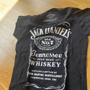 Jack Daniel's svart t-shirt, medium - Svart t-shirt från Jack Daniel's med ikoniskt vitt tryck framtill. Klassisk rund hals och korta ärmar. Tillverkad i 100% bomull för en skön och avslappnad känsla. Perfekt för dig som gillar rockig stil och vill sticka ut med en statement-tee.