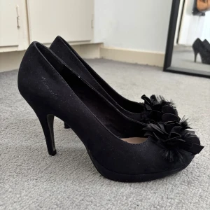 Svarta pumps från Tamaris med blomdetalj - Snygga svarta pumps från Tamaris med hög klack och rund tå i storlek 39. Köpte från Sellpy och säljer för att de är för stora för mig. De står att de är i nyskick🖤