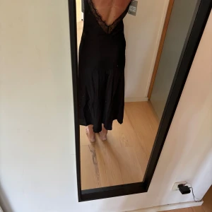 Svart långklänning med spetsdetalj - Svart långklänning i satinliknande material med djup v-ringning och öppen rygg. Klänningen har tunna axelband och spetskant längs ryggen. Aldrig använd